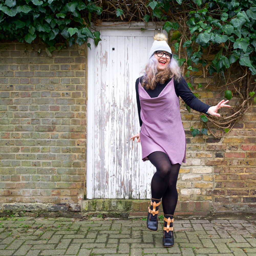 Janene is wearing her handmade Sicily slip dress and is skipping towards the lens in front on a white door surrounded by brickwork and ivy overhead. She is wearing a self-drafted black turtleneck top, grey bobble hat, black rimmed glasses and Vivienne Westwood Bondage Boots.