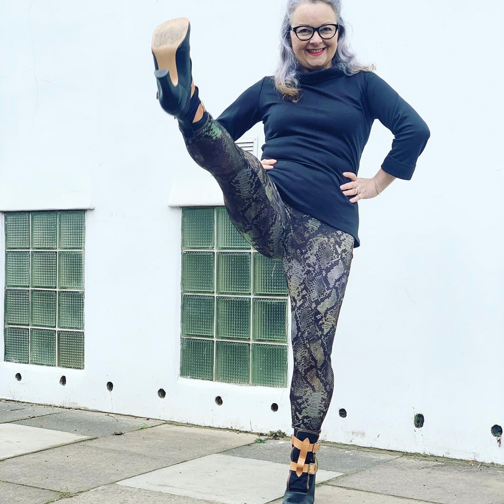 Image of Janene doing a high kick in her handmade snakeskin leggings. She is also wearing high heel boots, a black rollneck top and a pair of black rimmed glasses.
