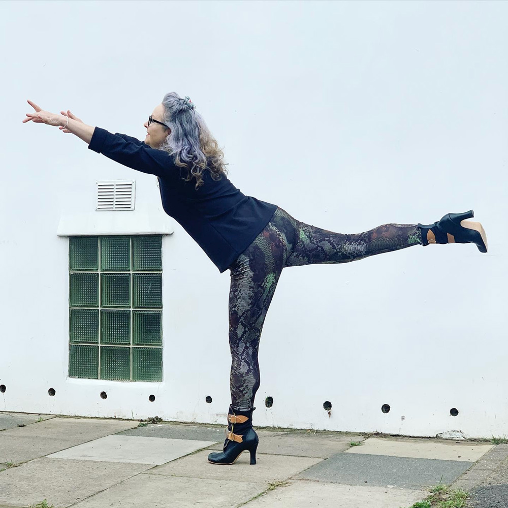 Janene is posing sideways, standing on one leg with the other leg straight out and arms forward. She is wearing her snakeskin print leggings with high heel boots and a black rollneck top