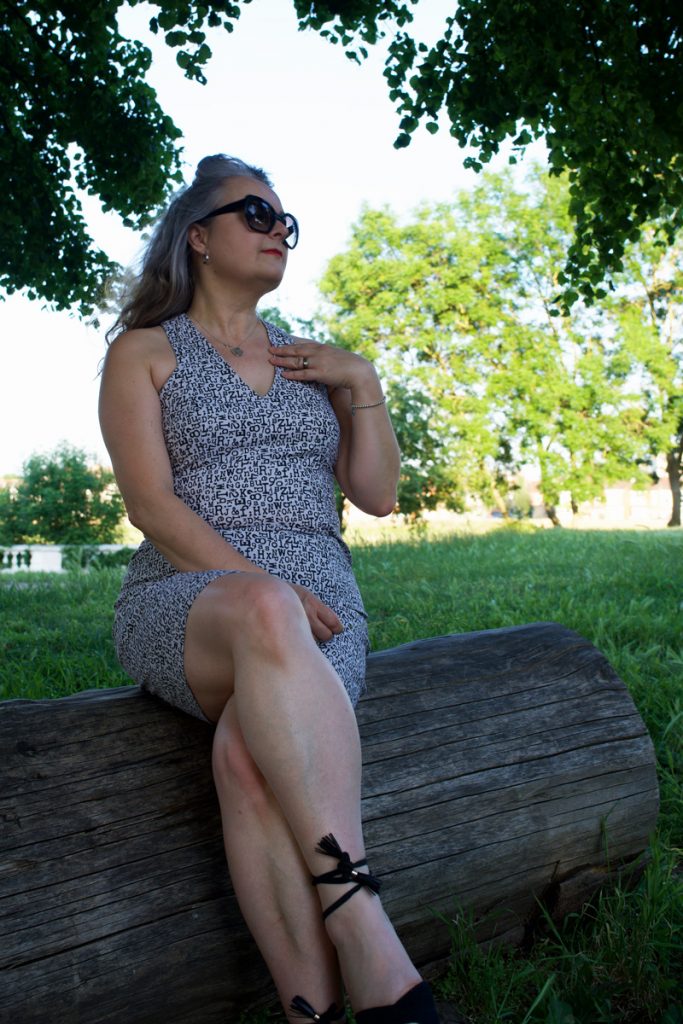 Janene is sitting on a log in the shade wearing her handmade dress, sunglasses and espadrilles. She is looking into the distance with her left hand on her shoulder and the other on her lap.