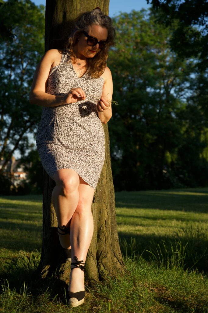 Janene is leaning against a tree holding a piece of grass, wearing her handmade sundress