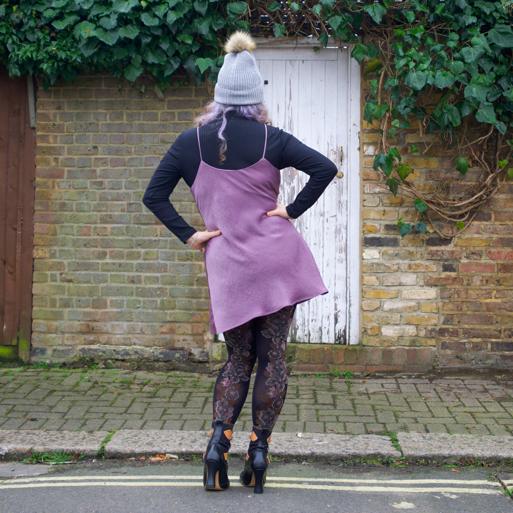 Back vew of Janene wearing her handmade Sicily slip dress. She is posing with both hands on hips in front on a white door surrounded by brickwork and ivy overhead. She is wearing a self-drafted black turtleneck top, grey bobble hat and Vivienne Westwood Bondage Boots.