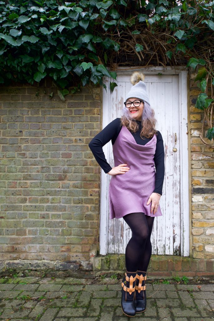 Janene is wearing her handmade Sicily slip dress and is posing with one hand on hip in front on a white door surrounded by brickwork and ivy overhead. She is wearing a self-drafted black turtleneck top, grey bobble hat, black rimmed glasses and Vivienne Westwood Bondage Boots.