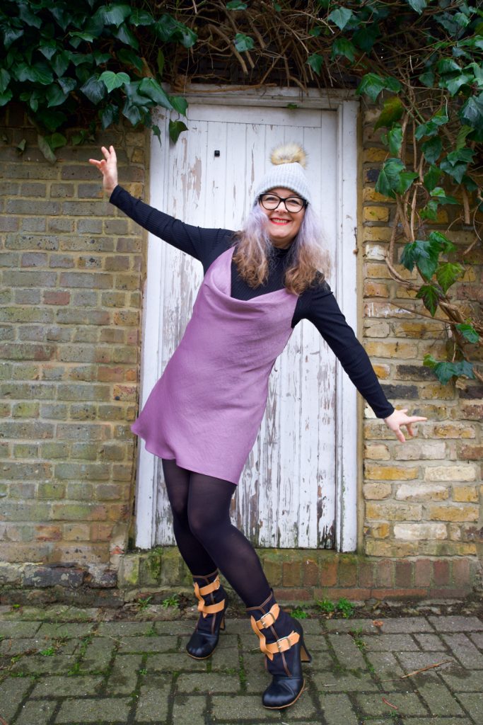 Janene is wearing her handmade Sicily slip dress and is posing with arms outstretched in front of a white door surrounded by brickwork and ivy overhead. She is wearing a self-drafted black turtleneck top, grey bobble hat, black rimmed glasses and Vivienne Westwood Bondage Boots.