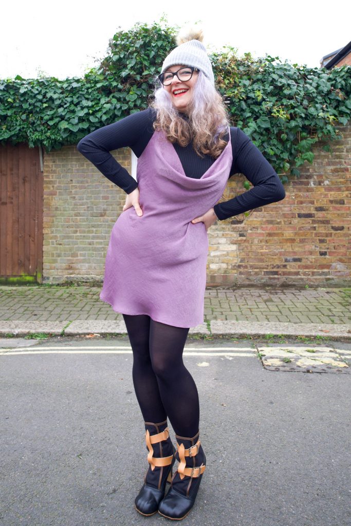 Janene is wearing her handmade Sicily slip dress and is posing with both hands on hips in front on a white door surrounded by brickwork and ivy overhead. She is wearing a self-drafted black turtleneck top, grey bobble hat, black rimmed glasses and Vivienne Westwood Bondage Boots.