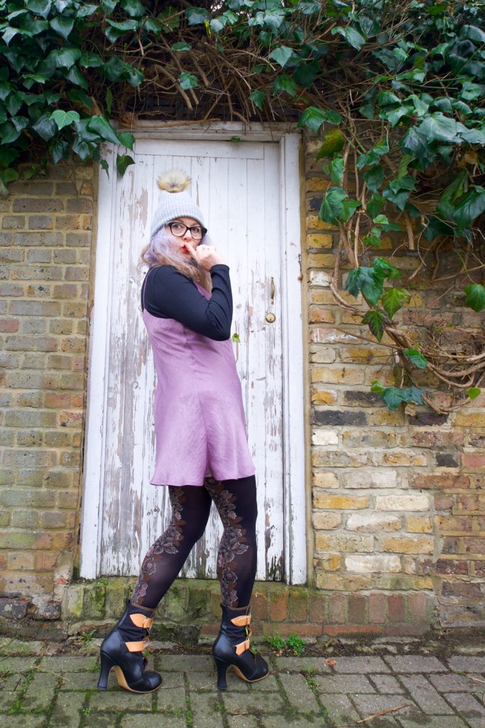 Side view of Janene wearing her handmade Sicily slip dress and is posing with right index finger on lip in front on a white door surrounded by brickwork and ivy overhead. She is wearing a self-drafted black turtleneck top, grey bobble hat, black rimmed glasses and Vivienne Westwood Bondage Boots.
