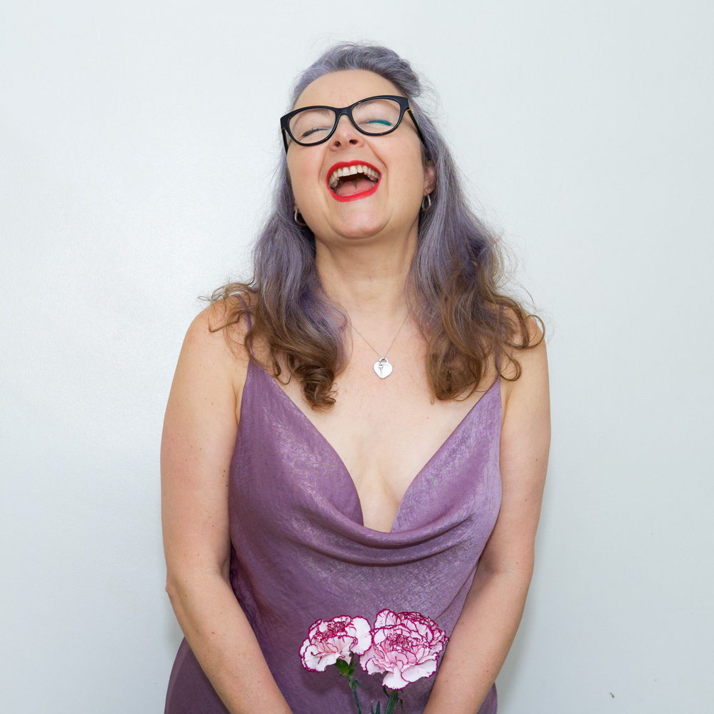 waist up image of Janene, wearing Masin Sicily slip dress. She is holding 3 lilac colour carnations with both hands to the bottom of the frame. She is wearing black rimmed glasses and her head is back, laughing.